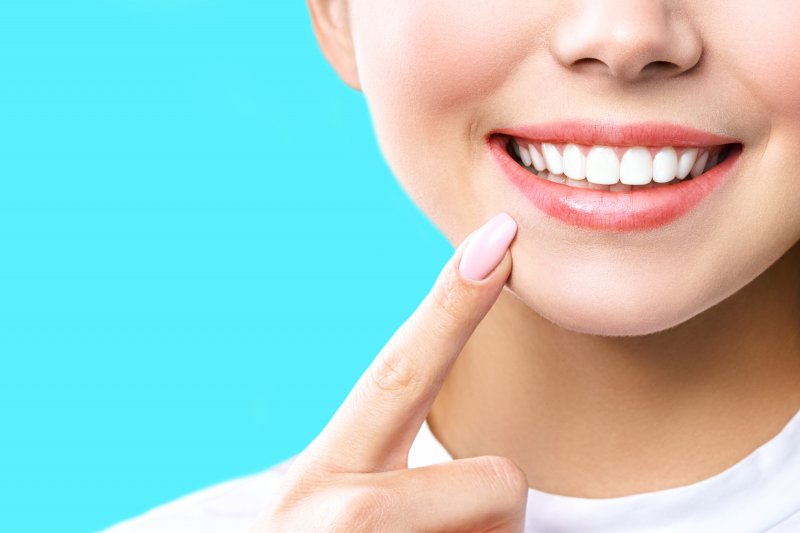 Woman in white tee pointing index finger at her whitened teeth on teal background